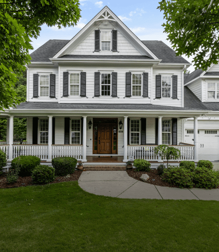 Front view of a white two-story house with dark shutters, a wooden front door, a wide porch, and an attached garage. The yard features green grass, bushes, and trees, with a walkway leading to the porch.