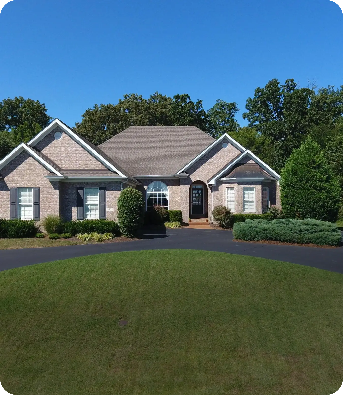 A brick house with a central entrance, multiple windows, and a landscaped front yard with a circular driveway on a clear sunny day. Trees are in the background.
